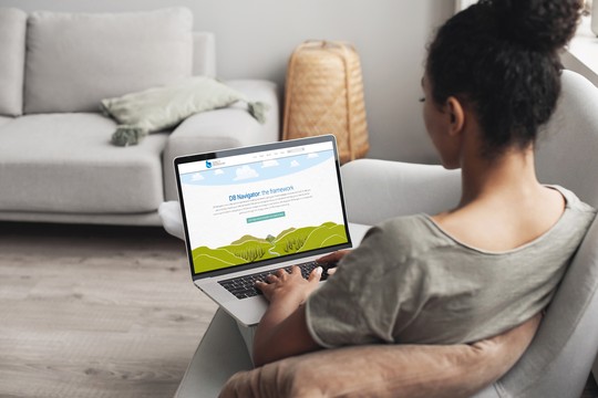 A person sits on a couch, using a laptop displaying the "DB Navigator: the framework" webpage. The cozy room features soft furnishings and a warm atmosphere, enhancing the relaxed setting.