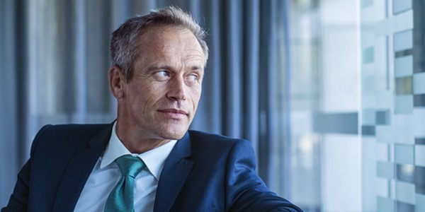 A man in a dark suit and light blue tie rests his arm on a glass surface, gazing thoughtfully out of a large window, surrounded by softly lit curtains and a modern interior.