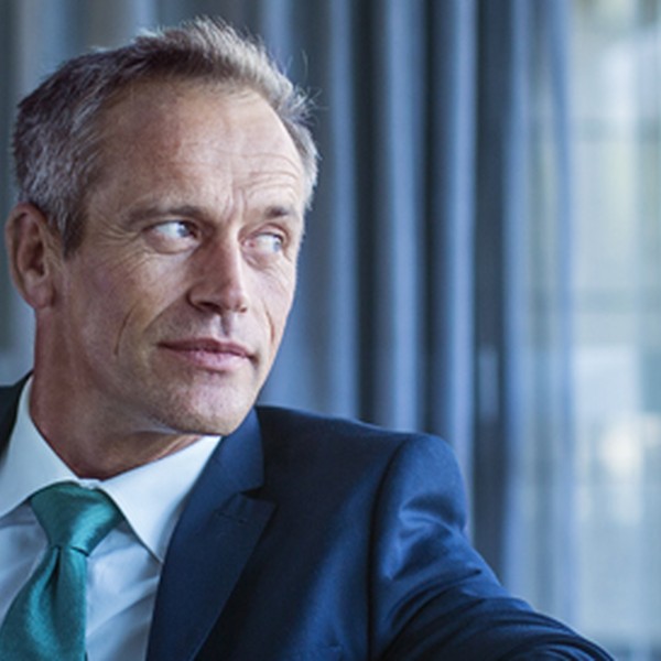 A man in a dark suit and light blue tie rests his arm on a glass surface, gazing thoughtfully out of a large window, surrounded by softly lit curtains and a modern interior.