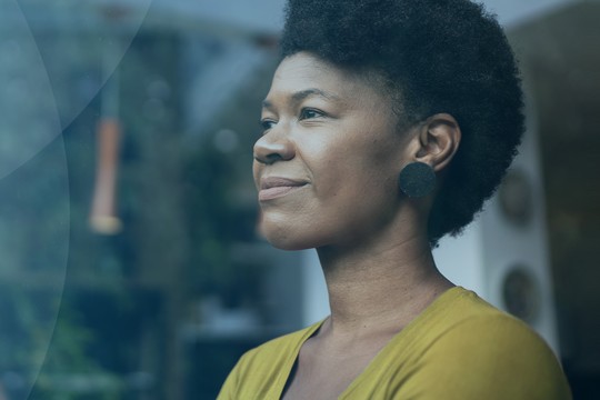 A woman with natural hair is gazing thoughtfully out of a window. The setting is bright with greenery visible outside, suggesting a serene and contemplative atmosphere.