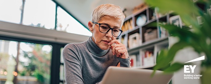 A woman with short, light-colored hair and glasses is concentrating on her laptop, resting her chin on her hand. She is in a bright room with bookshelves and plants.