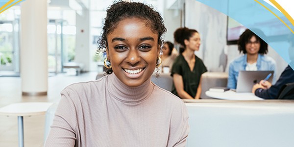 A young woman with curly hair smiles at the camera while seated at a table. In the background, several individuals are engaged in conversation and working on laptops in a modern, bright setting.