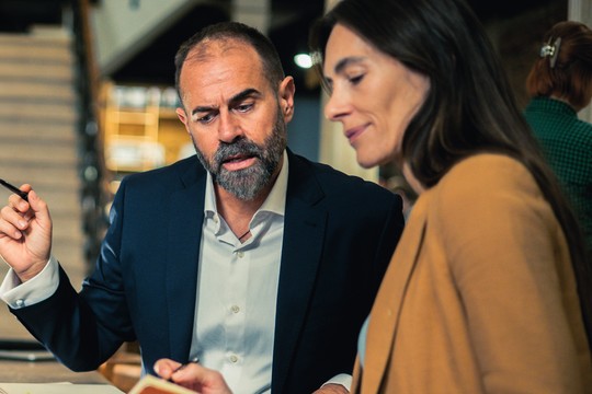 A man in a suit gestures animatedly while discussing something with a woman in a brown jacket, both focused on a notebook between them in a casual office setting.