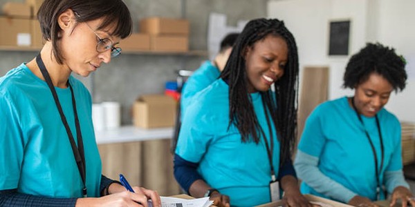 Three women in teal shirts are working in a warehouse, where one is writing on a form, while the others smile and open cardboard boxes. The background features shelves and packaging materials.