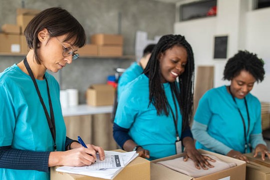 Three women in teal shirts are working in a warehouse, where one is writing on a form, while the others smile and open cardboard boxes. The background features shelves and packaging materials.