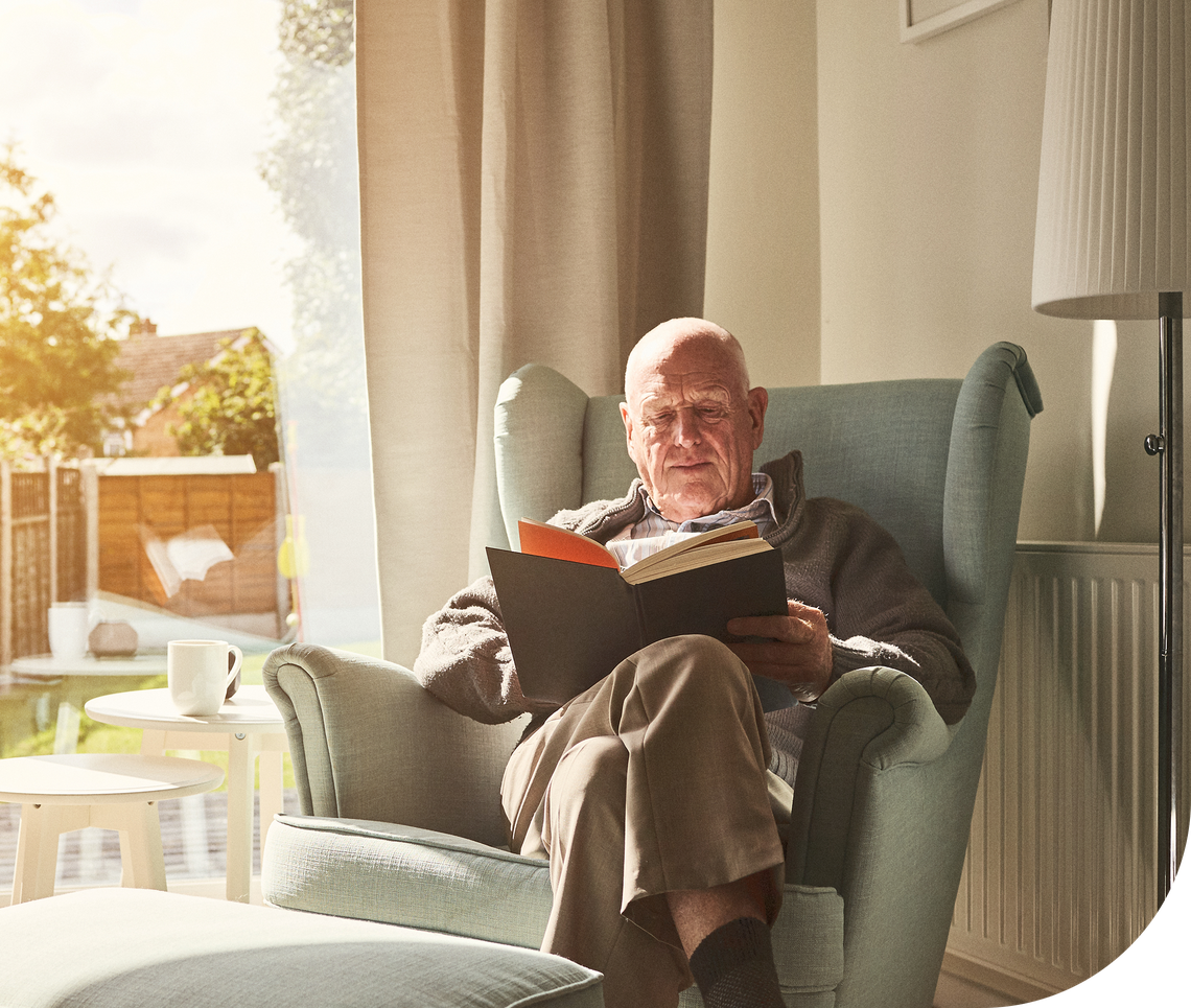 An elderly man sits comfortably in a light blue armchair, reading a book. Sunlight streams through a window, illuminating a cozy living space with a small table and a cup nearby.