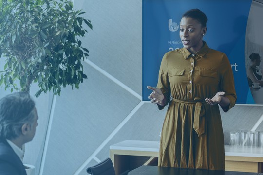A woman stands and speaks in a conference room, engaging with two seated men. A large screen displays a meeting backdrop, and a plant is visible nearby, adding to the professional setting.