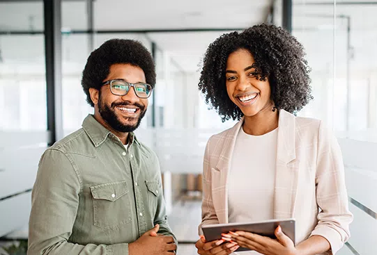 Two smiling professionals stand close together in a modern office space, exchanging positive energy. One holds a tablet, while the other crosses their arms, both appearing engaged and friendly.