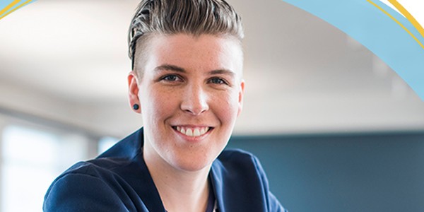 A person with short hair and a blue blazer smiles confidently, leaning on a table in a bright, modern office environment with large windows and a neutral backdrop.