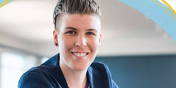 A person with short hair and a blue blazer smiles confidently, leaning on a table in a bright, modern office environment with large windows and a neutral backdrop.