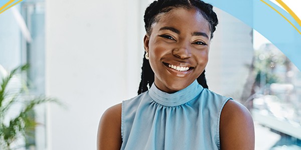 A woman with braided hair smiles warmly, wearing a sleeveless blue top. She stands indoors, facing the camera, with a bright environment and greenery visible in the background.