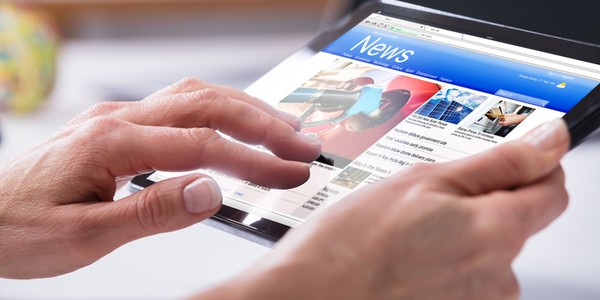 A hand interacts with a tablet displaying a news website. The screen shows articles and images, set against a blurred indoor background with soft lighting.