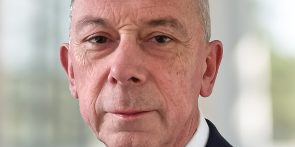 A middle-aged man in a dark suit and tie stands facing the camera, displaying a serious expression, with a softly blurred professional background suggesting an office or corporate setting.