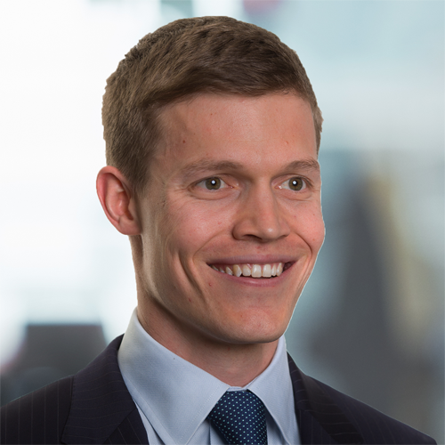 A young man with short, light brown hair smiles, dressed in a dark suit and tie. He is in a professional setting, with a blurred background suggesting an office environment.