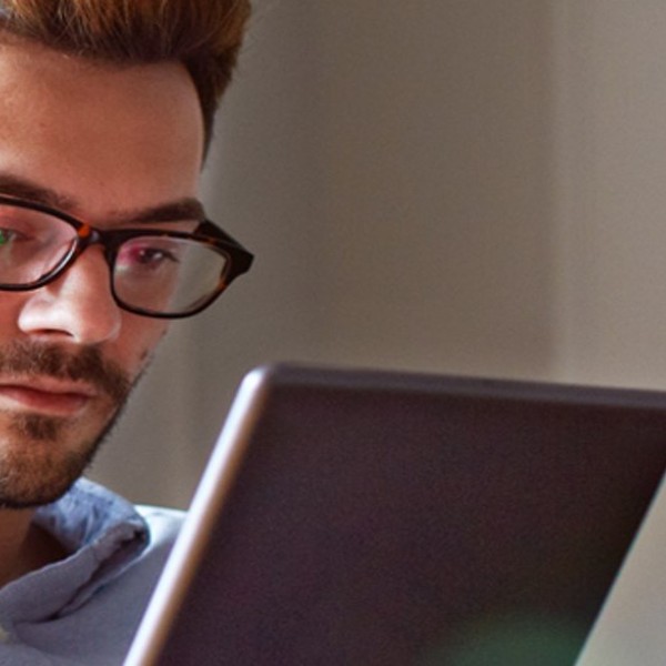 A man with glasses is focused on a tablet, sitting comfortably in a casual outfit, with a softly lit interior background, suggesting a relaxed setting for work or leisure.