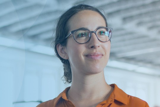 A woman in an orange shirt and glasses stands, smiling gently. She appears engaged in conversation within a bright, modern office environment with a blurred background.