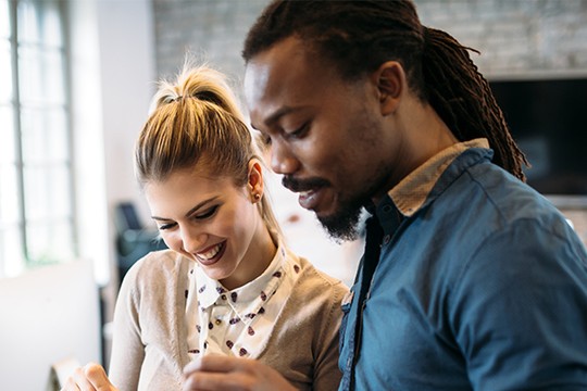 A woman and a man stand closely together, smiling as they examine something on a device. The setting features a modern, well-lit workspace with a brick wall backdrop.