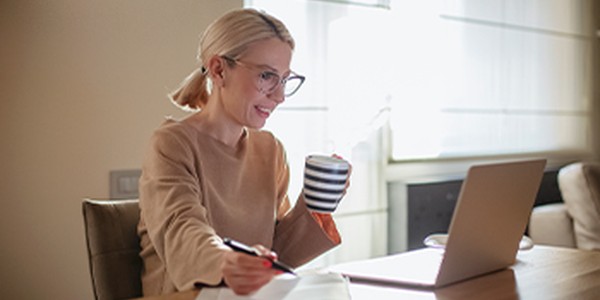 A woman with glasses sits at a table, holding a striped mug and writing notes, while smiling at her laptop. Sunlight filters through sheer curtains, creating a warm, cozy atmosphere.