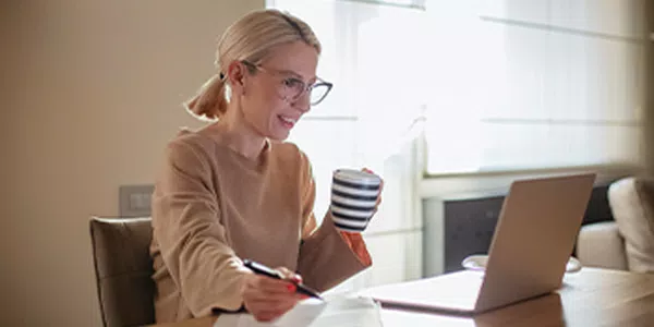 A woman with glasses sits at a table, holding a striped mug and writing notes, while smiling at her laptop. Sunlight filters through sheer curtains, creating a warm, cozy atmosphere.