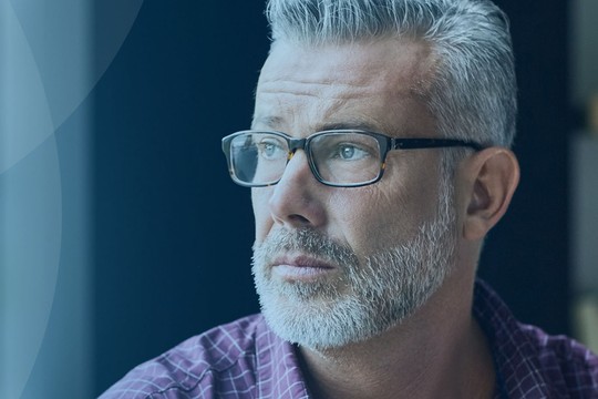 A man with glasses and gray hair gazes thoughtfully out a window, seated in a softly lit room with shelves and a hint of books in the background.