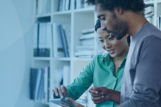 Two individuals interact while looking at a tablet together in a modern office. The woman, wearing a green shirt, smiles, and the man in a gray shirt points at the screen, suggesting collaboration.