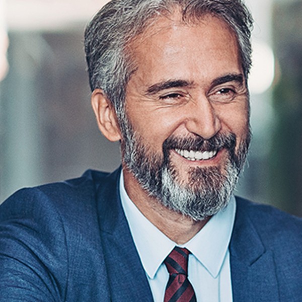 A middle-aged man in a blue suit is smiling and engaging with two others during a meeting in a modern office setting, which features glass walls and soft lighting.