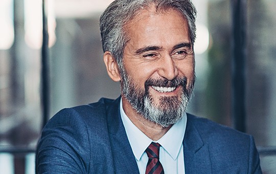 A middle-aged man in a blue suit is smiling and engaging with two others during a meeting in a modern office setting, which features glass walls and soft lighting.