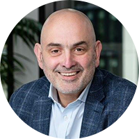A bald man with a beard and a friendly smile is seated indoors, wearing a light blue shirt and a dark blazer, with indoor plants and large windows in the background.