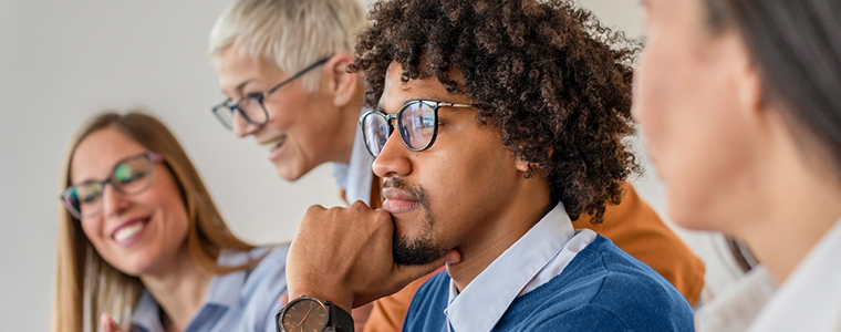 A man with curly hair sits thoughtfully, resting his chin on his hand, while three smiling women in glasses engage with one another in a bright, modern meeting space.
