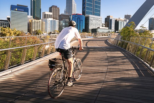 A cyclist rides on a wooden pathway over a bridge, surrounded by modern buildings and trees, with clear skies and a vibrant cityscape in the background.