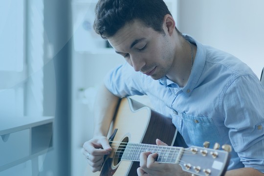 A young man sits in a black office chair, playing an acoustic guitar while focused on his music. A laptop is visible on the desk in a bright, modern room.