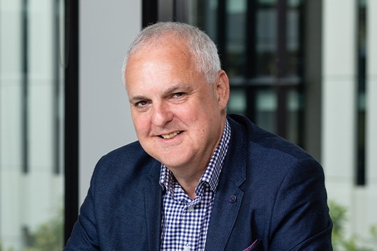 A man with short gray hair smiles warmly while sitting at a table. He wears a blue blazer over a checkered shirt, with a modern office environment visible through large windows behind him.