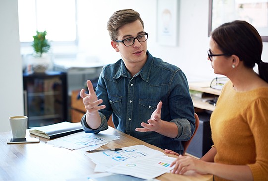 A young man in a denim shirt gestures as he discusses documents with a woman in a yellow sweater. They sit at a table surrounded by papers and a coffee cup, in a bright office setting.