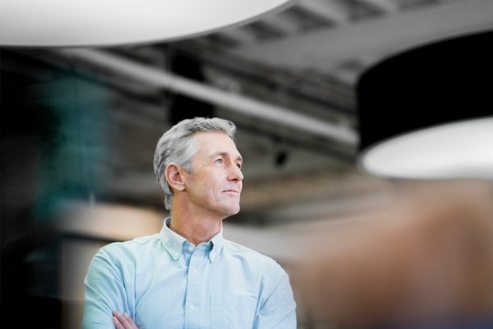 A mature man stands confidently with his arms crossed, looking thoughtfully upward in a modern, well-lit office space featuring suspended lighting fixtures and a contemporary design.