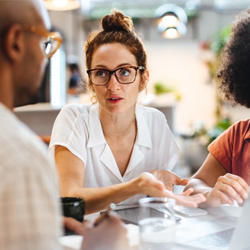 Three individuals are engaged in a discussion around a table. One person gestures while speaking. They are in a bright, modern workspace with plants in the background and a laptop in front of them.