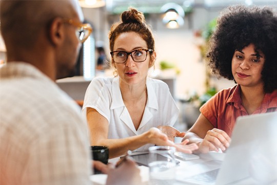 Three individuals are engaged in a discussion around a table. One person gestures while speaking. They are in a bright, modern workspace with plants in the background and a laptop in front of them.