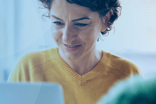 A person with curly hair, wearing a yellow sweater, smiles as they look at a laptop, holding a pair of glasses. The setting appears bright and modern, with soft colors.