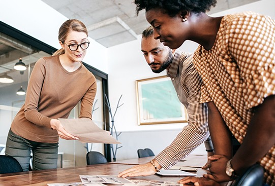 A woman in glasses points at papers while discussing them with two colleagues. They are in a modern office with large windows, wooden furniture, and artistic decor on the walls.