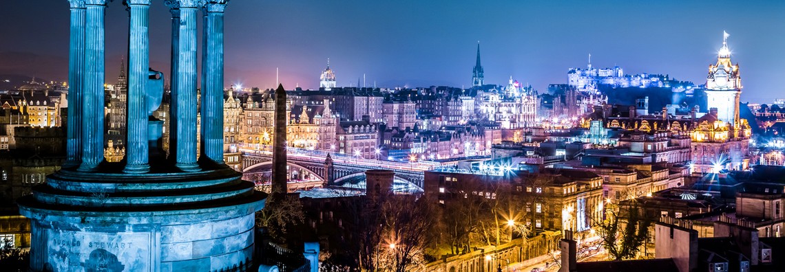A tall monument with columns stands prominently in the foreground, while a vibrant cityscape of Edinburgh glows at night, filled with historic buildings and a castle in the background.