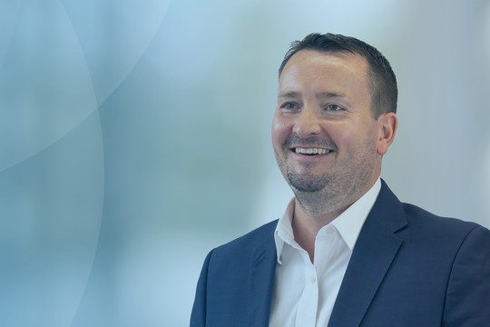 A man in a dark suit smiles, standing against a blurred light blue background with soft curves. He appears engaged and approachable, suggesting a professional context.