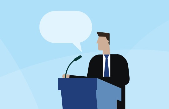 A man in a suit stands at a podium with a microphone, delivering a speech. He faces an audience in a light blue environment, indicated by a speech bubble above him.