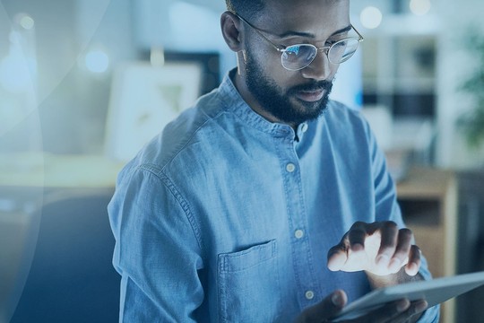 A man in glasses interacts with a tablet, focused on the screen. He is seated in a dimly lit room, with soft lighting and plants in the background, suggesting a cozy environment.