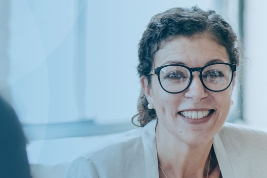 A woman with curly hair and glasses smiles while engaging in conversation with others in a bright, modern meeting space, conveying a positive and collaborative atmosphere.