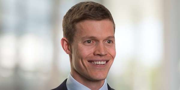 A young man with light brown hair smiles confidently, wearing a dark suit and light blue shirt. The soft-focus background suggests an indoor setting, possibly an office or corporate space.