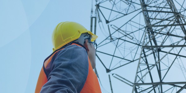 A worker in a hard hat and safety vest examines a tall electrical tower against a clear blue sky, indicating attention to safety and infrastructure inspection.