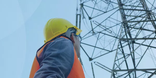 A worker in a hard hat and safety vest examines a tall electrical tower against a clear blue sky, indicating attention to safety and infrastructure inspection.