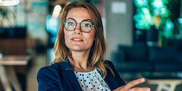 A woman wearing glasses and a blazer gestures while speaking, her expression thoughtful. She is seated in a modern, indoor space with soft lighting and a hint of greenery in the background.