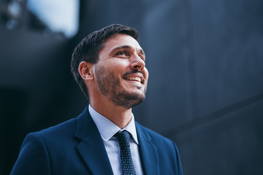 A man in a suit is smiling and looking upwards. He stands in a modern urban environment, with dark, textured walls in the background suggesting a contemporary architectural setting.