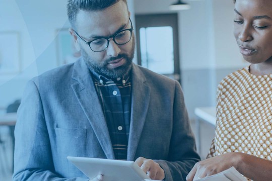 Two professionals engage collaboratively, one using a tablet to display information. They are in a modern, bright office space with an open layout and large windows.