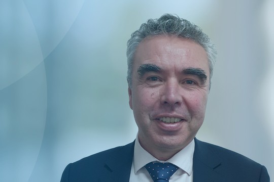 A man in a suit smiles warmly, captured against a soft blue gradient background. The setting appears professional, suggesting an office or corporate environment.